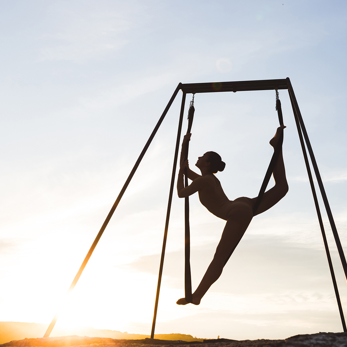 Woman using pole aerial silks to perform fly yoga stretch at sunset